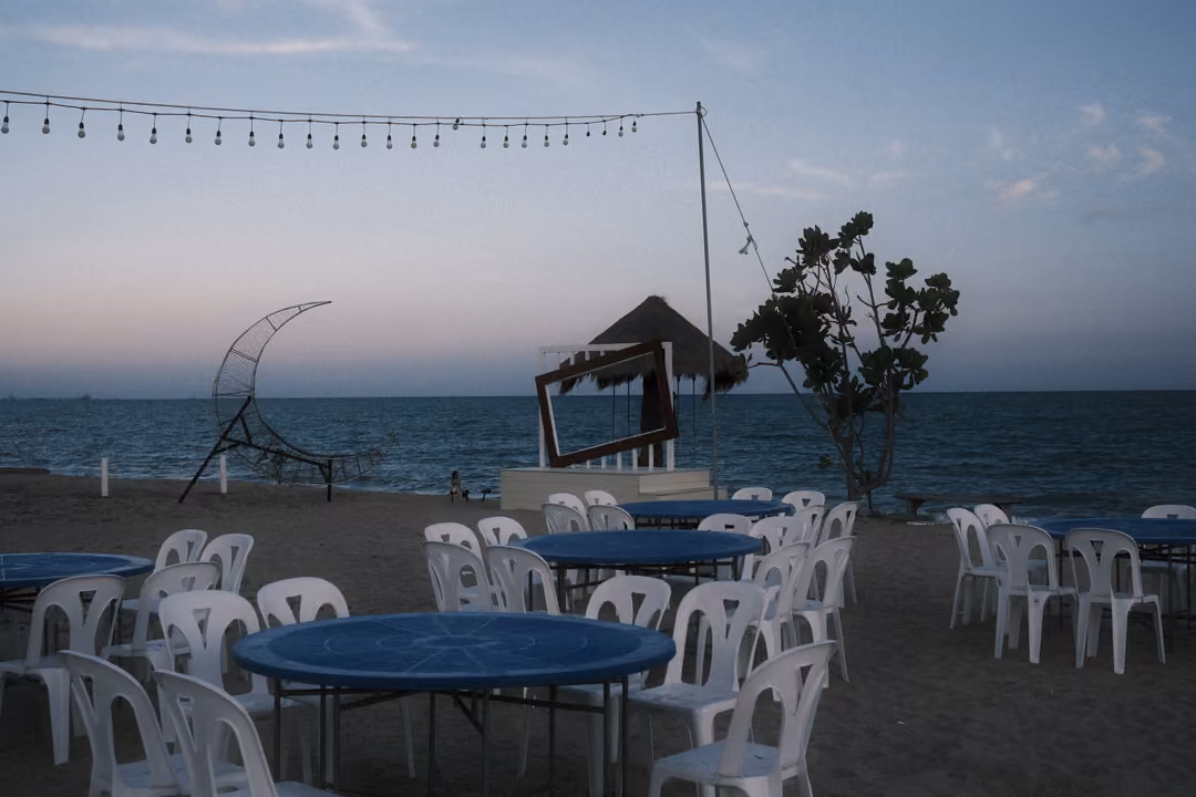 Beachfront with white plastic chairs around blue tables, string lights overhead and the sea at dusk