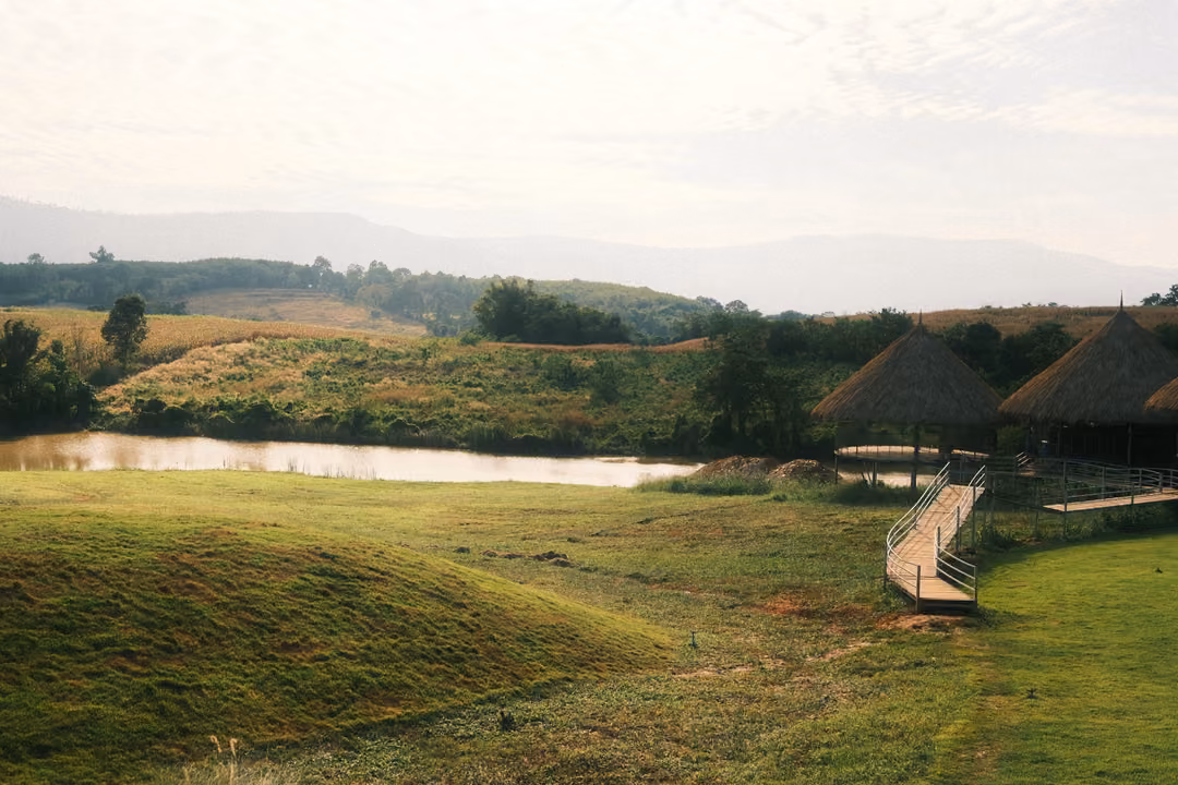 Grassy mounds lead down to a pond, with a wooden ramp climbing to thatched huts on the right.