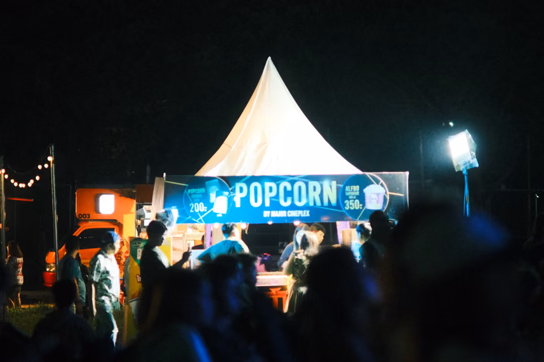 Crowd queuing at a lit popcorn stand under a white peaked tent at night, string lights at left.