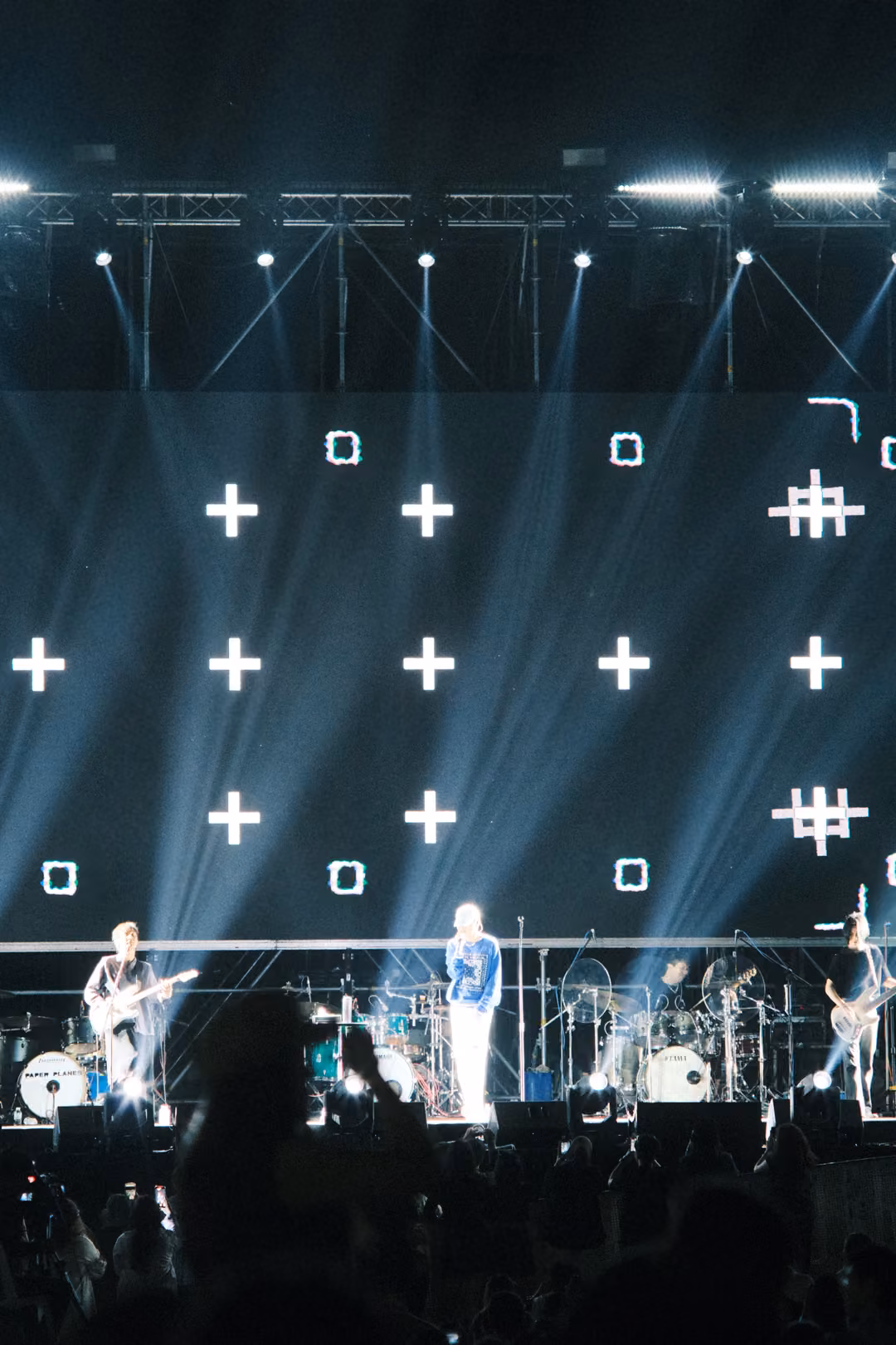 Vertical view of a screen of white crosses and hash marks above the singer and guitarists, a dark head in the foreground.