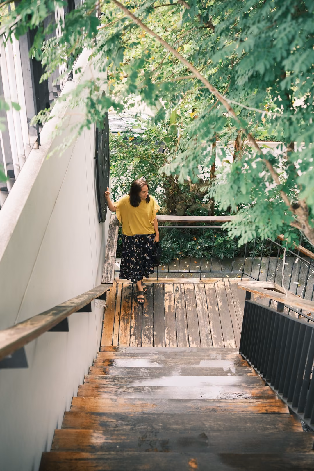A person in a yellow top stands at the top of a wet wooden staircase, hand on the wall, under leafy branches.