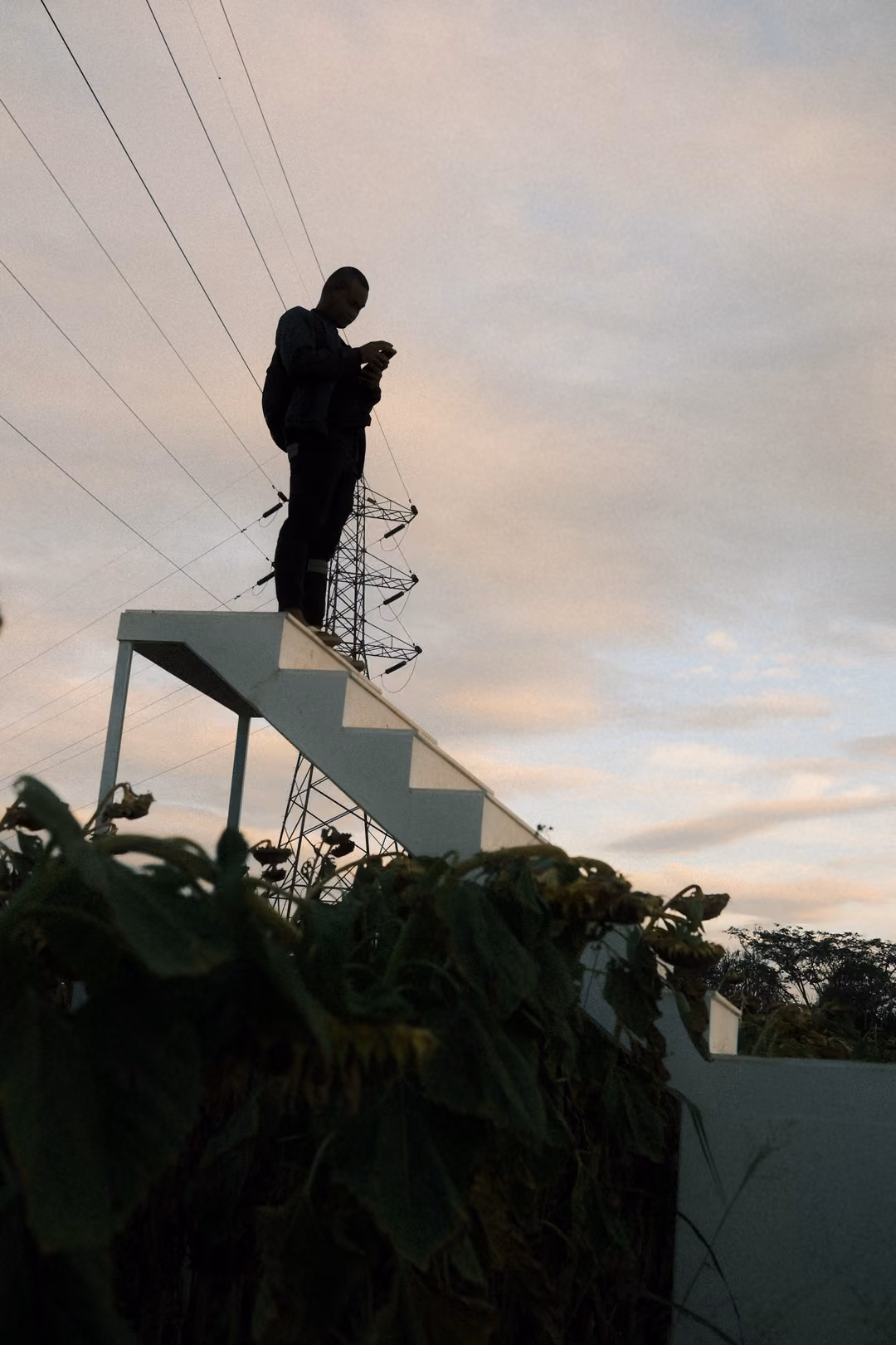 Silhouette of a person standing at the top of a white staircase beside a power pylon under a cloudy evening sky