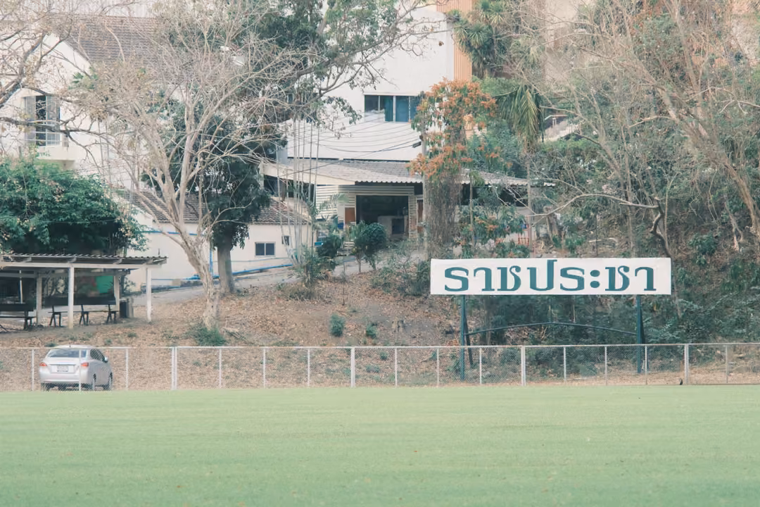 Green field in front of a fence and white sign with Thai lettering, a building and parked car behind