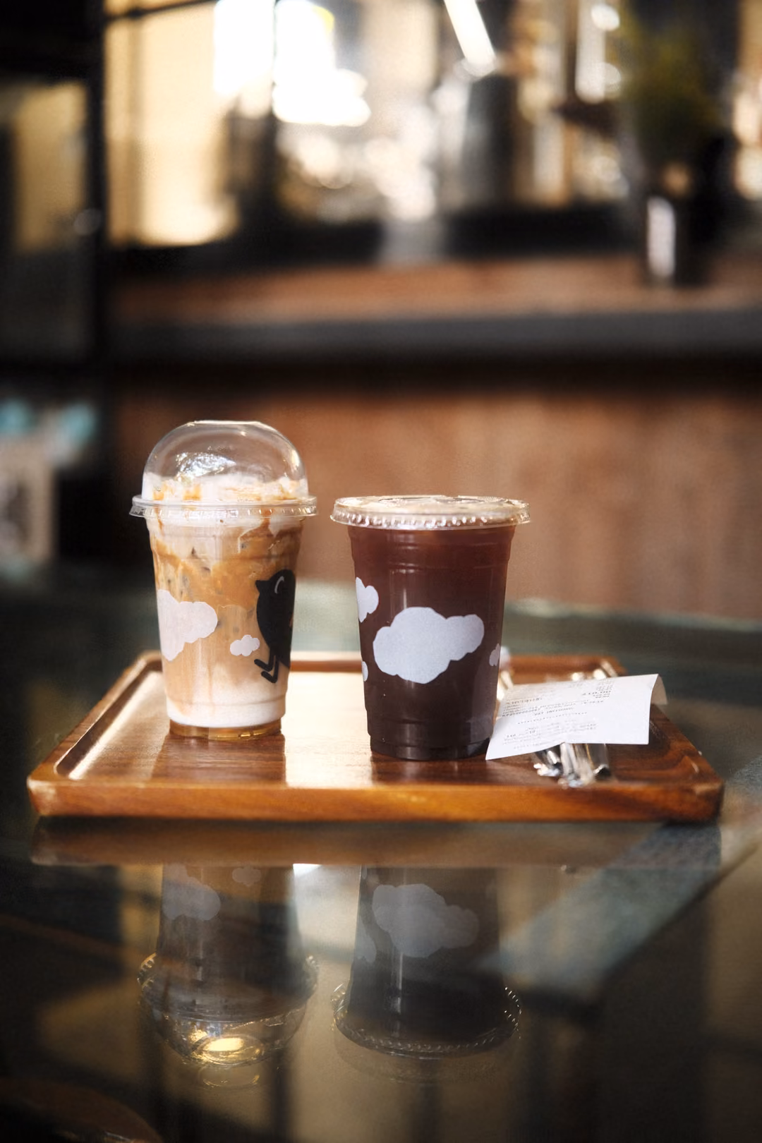 Two iced drinks in plastic cups with cloud prints on a wooden tray, reflected in a glass table