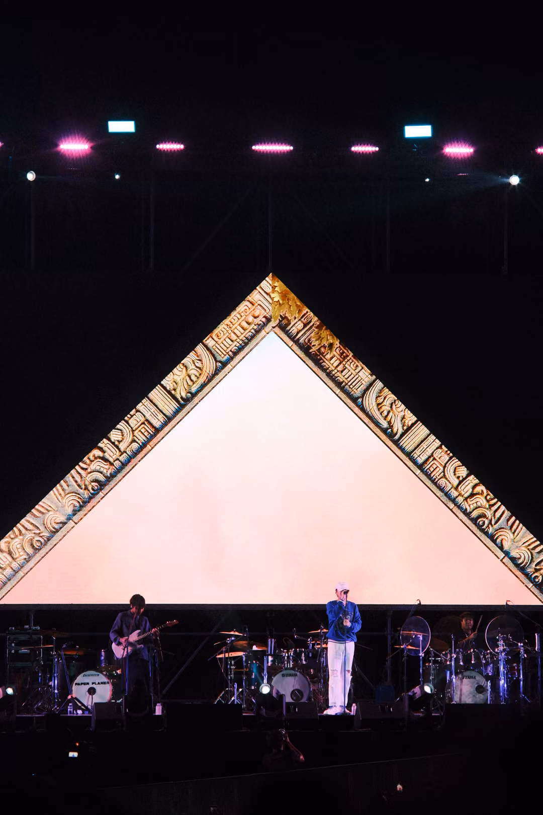 Vertical view of a gold-framed triangle glowing pale pink, the singer in a blue jacket and a guitarist below.