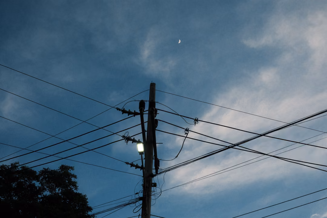 Power pole with a lit lamp and many wires against a blue sky, a faint crescent moon above, trees at left