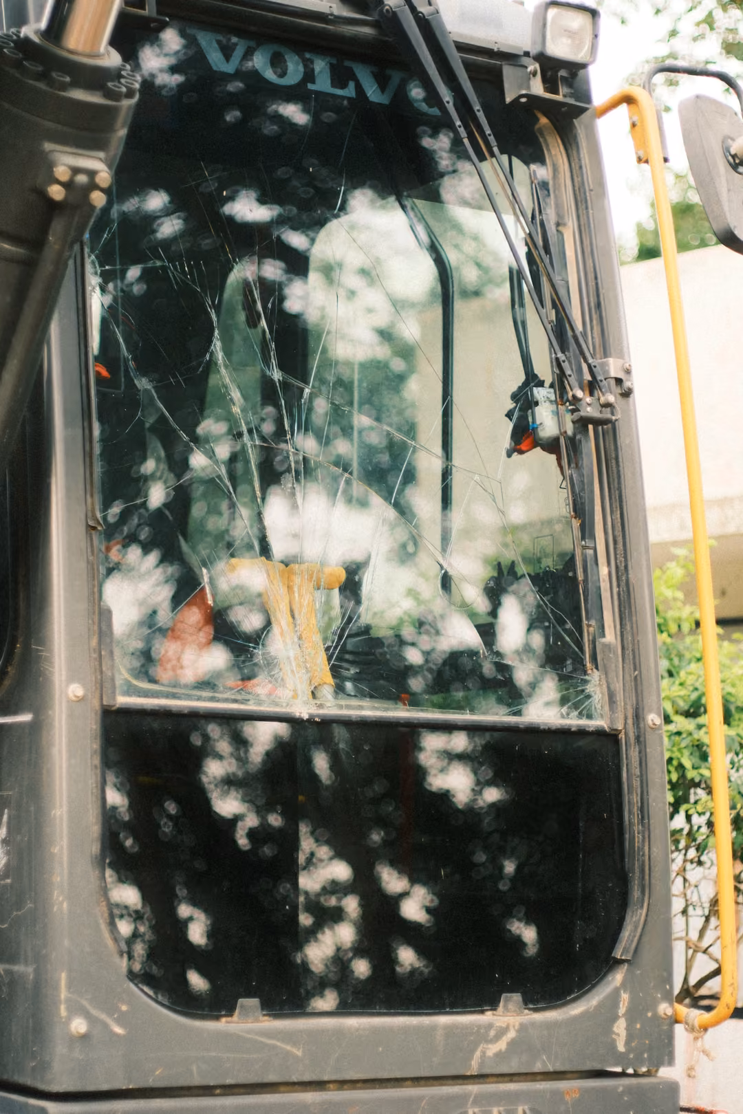 The cracked windscreen of a Volvo excavator cab, with tree reflections and a yellow handrail at the side.