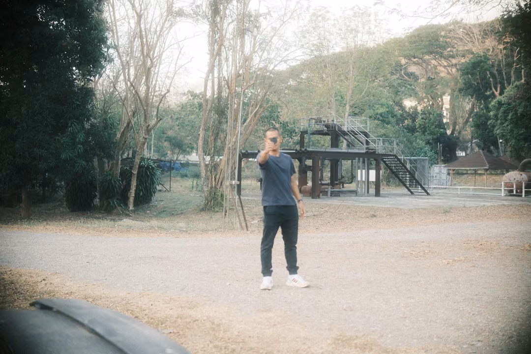 Man standing in a gravel yard holding a phone up in front of his face, metal structures and trees behind