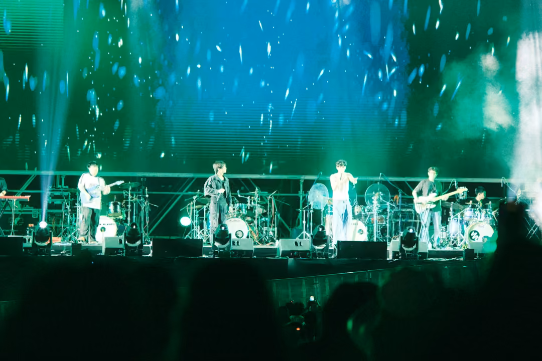 Band performing beneath a green and blue screen covered in falling white flakes, crowd heads silhouetted.