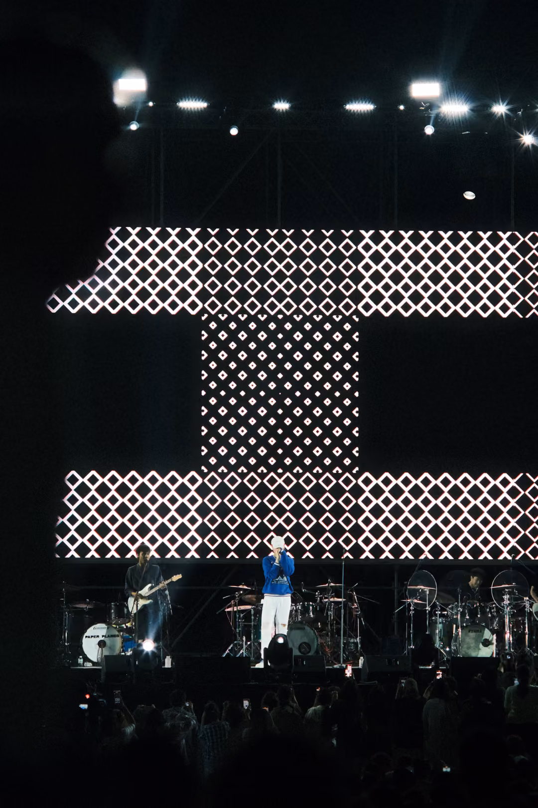 Vertical view of a screen with bands of white diamond patterns separated by black, singer and guitarist on stage below.