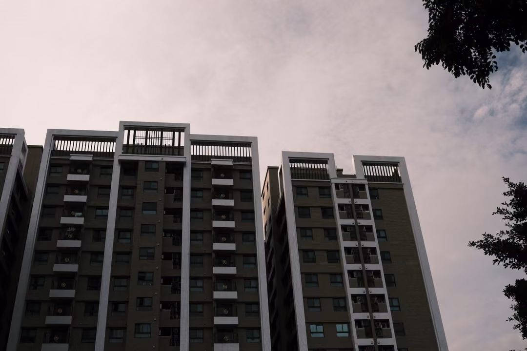 Low view looking up at tall residential towers with white frames against an overcast sky, tree edges at the side
