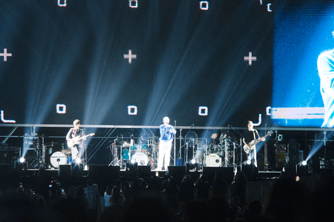 Stage with white plus signs and squares on screen, a blue panel showing a figure at right, band performing below.