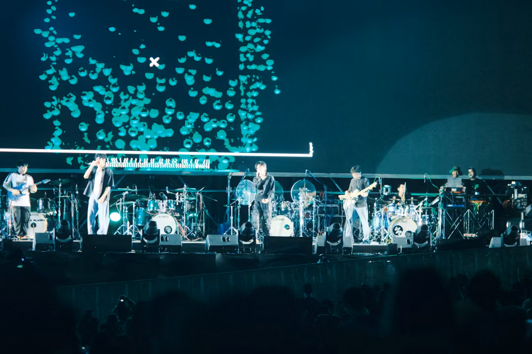 Band on stage beneath a dense cloud of turquoise graphic shapes on a large screen, crowd silhouetted in the foreground.