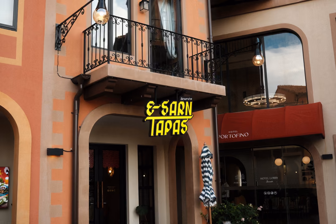 Yellow neon sign reading E-Sarn Tapas below a wrought iron balcony, beside a red Portofino awning