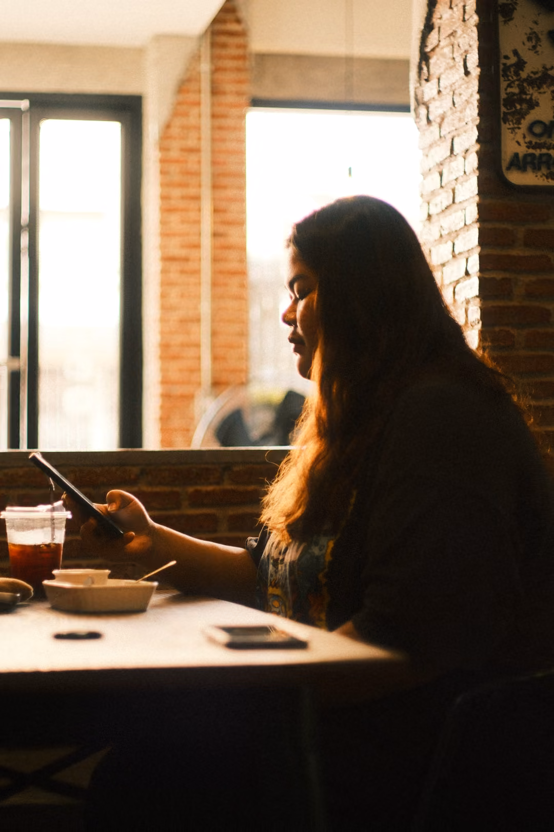 A woman with long hair looks down at her phone at a cafe table, backlit by a bright window