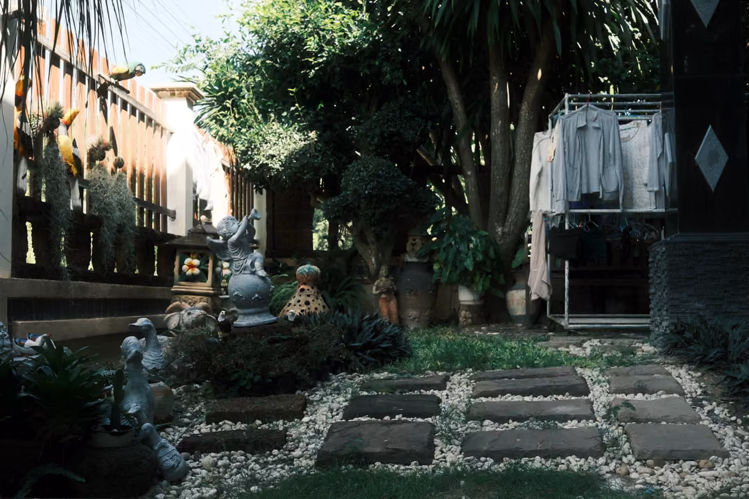Garden path of square stepping stones in white gravel, clothes on a rack and statues among palms