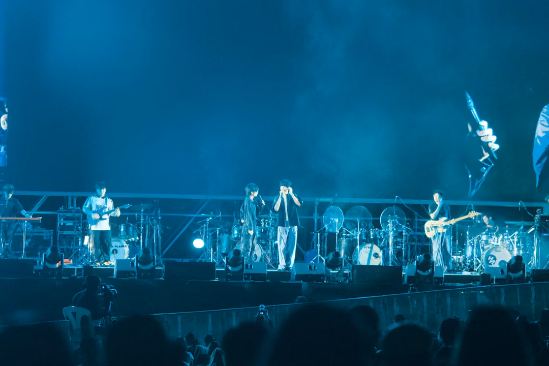 Band on a stage filled with teal haze, the singer holding a microphone at centre, a guitarist on each side.