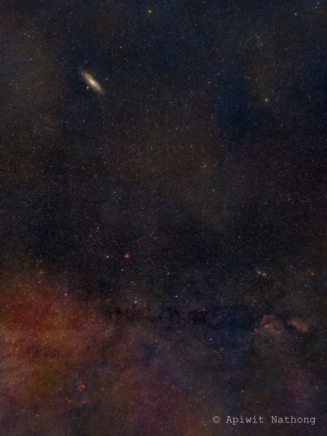 Tall starfield with a small elongated galaxy near the top and reddish clouds of the Milky Way below