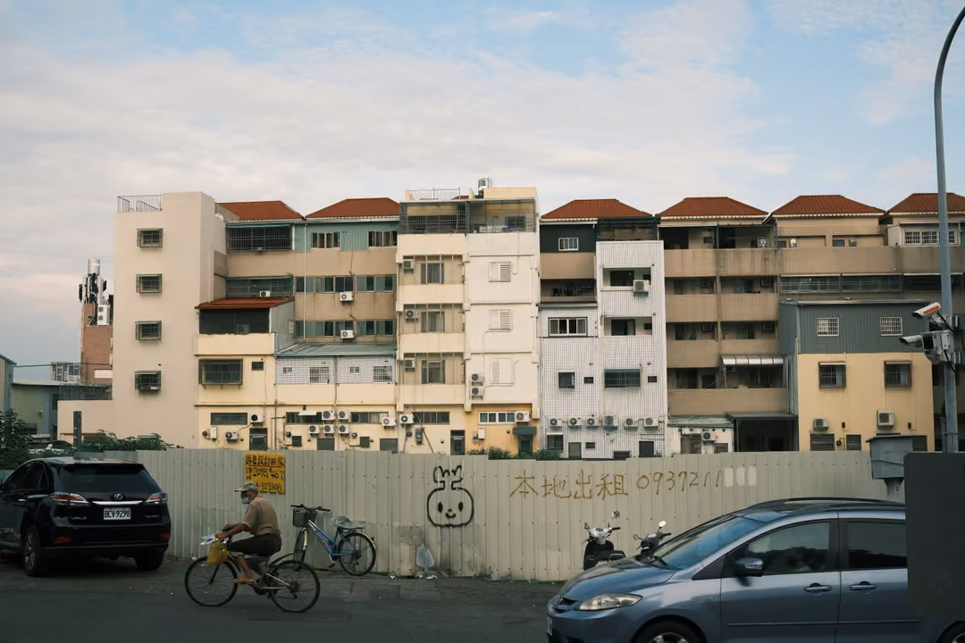 Street with an apartment block behind a construction fence, a cyclist riding past and parked cars