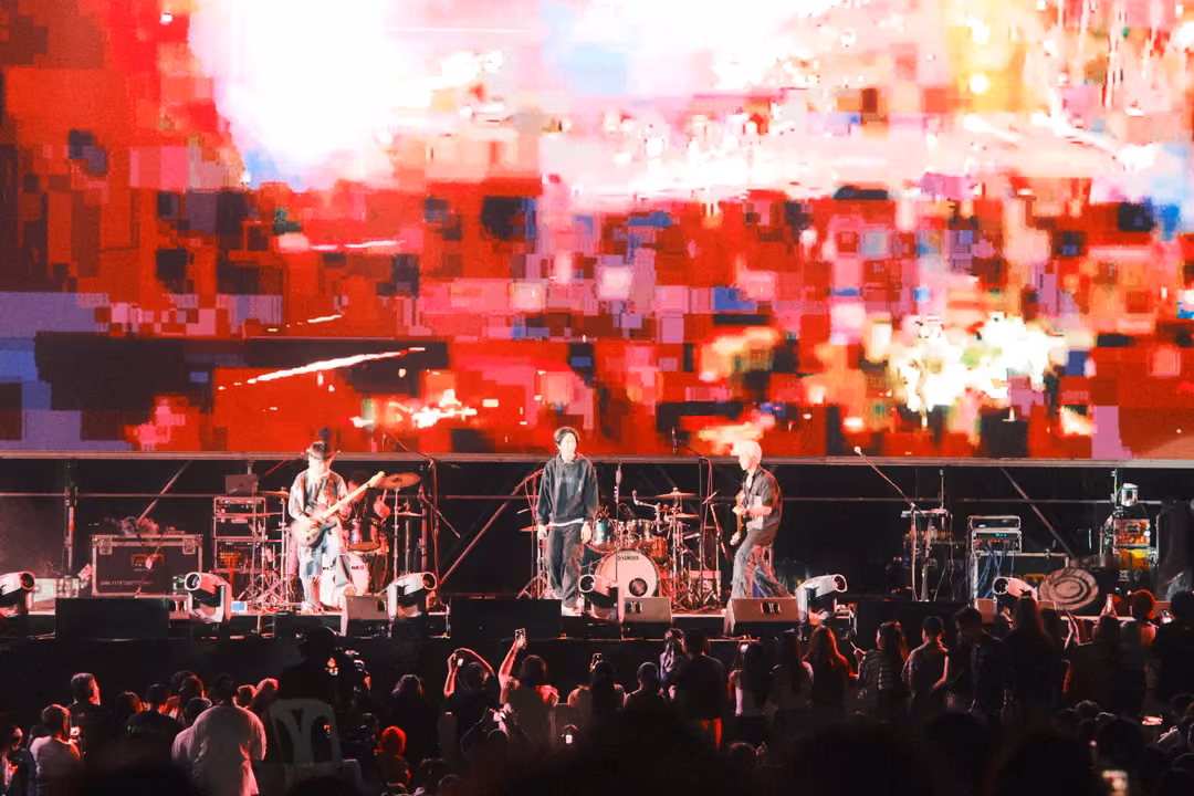 Band performing below a screen of glitched red and white pixel blocks, crowd with raised arms in the foreground.