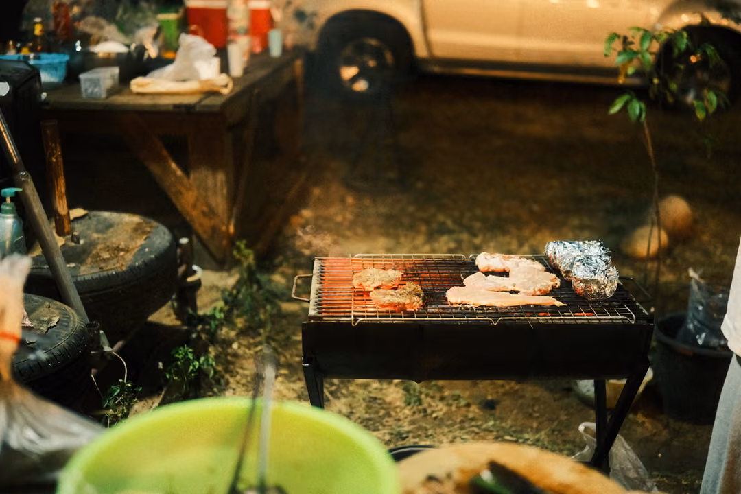 Grill with meat and foil packets beside a table of drinks and a tyre, a parked white car behind