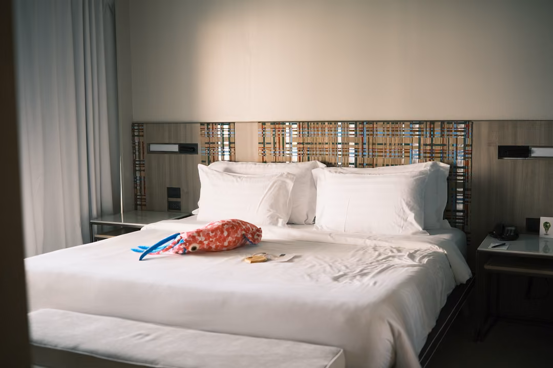 A hotel bed with white pillows and a woven headboard, a red bag and a blue item on the covers