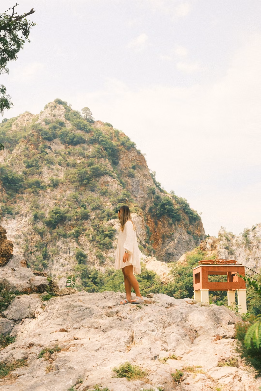 Woman in a white dress standing in profile on a rock ledge, looking out, hill and red sign box behind