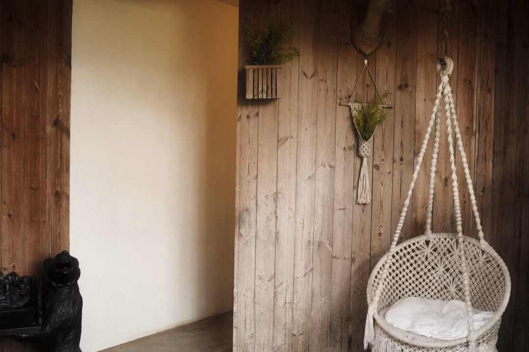 A white macramé hanging chair with a cushion against a wooden wall, a small plant hanger beside it