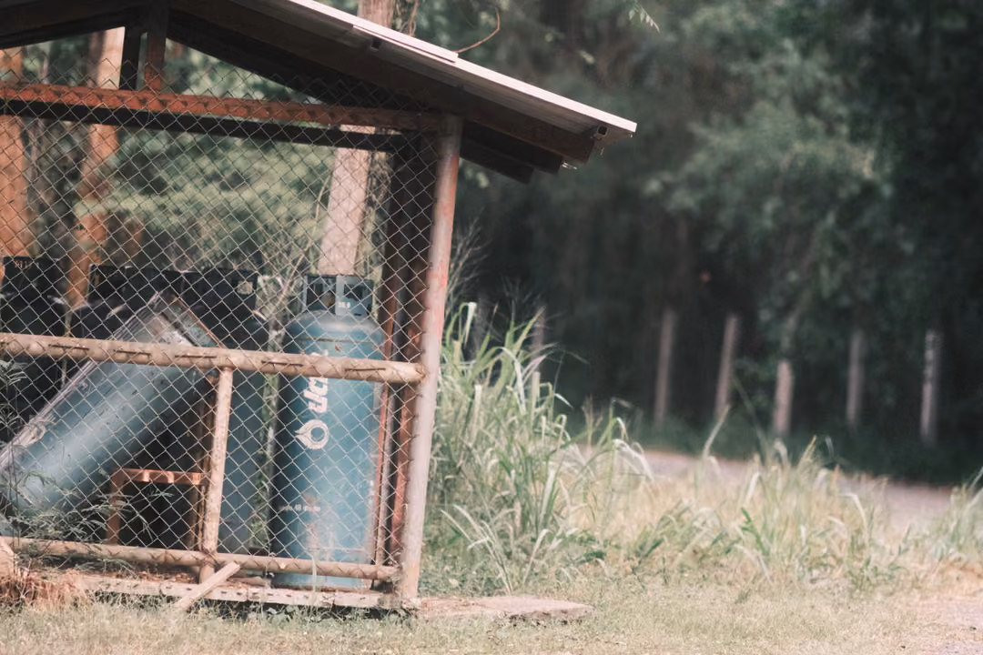Two green gas cylinders inside a fenced wire cage under a small roof, grass and a fence line nearby