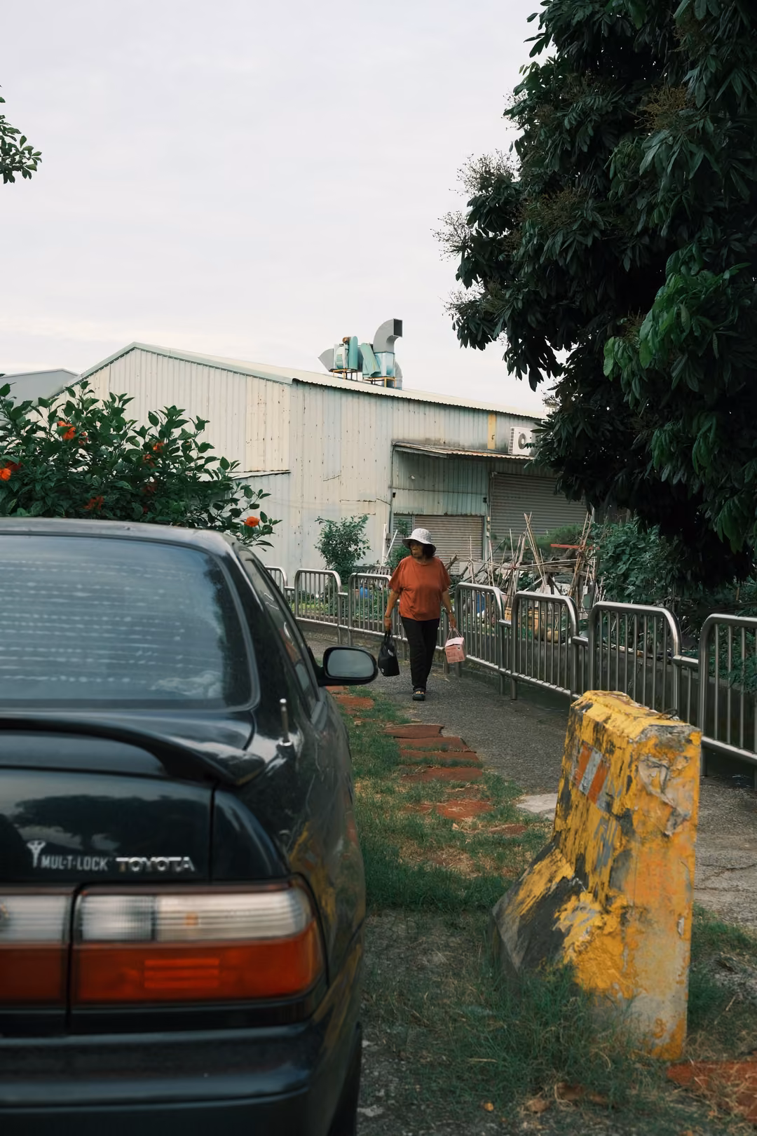 Rear of a dark Toyota sedan parked beside a grassy verge, a yellow bollard and a person walking past a fence