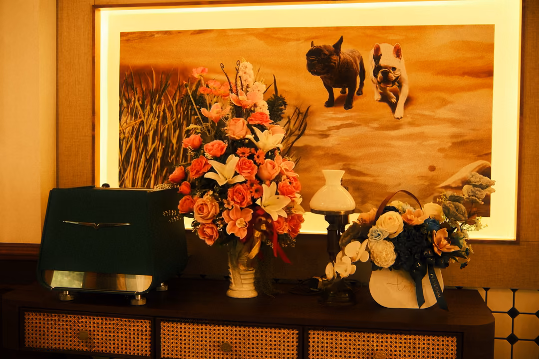 Backlit painting of two small dogs on sand above a cabinet with flower arrangements, a lamp and a green radio