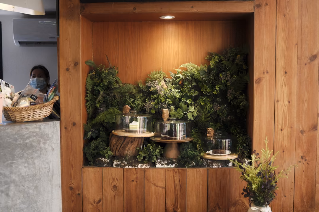 A wooden alcove filled with plants and glass cake domes on stands, a masked person at a counter