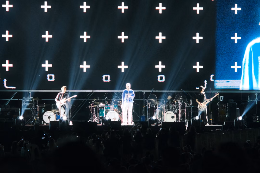 Band on stage under a screen grid of white plus signs, a blue panel at right showing part of the singer.
