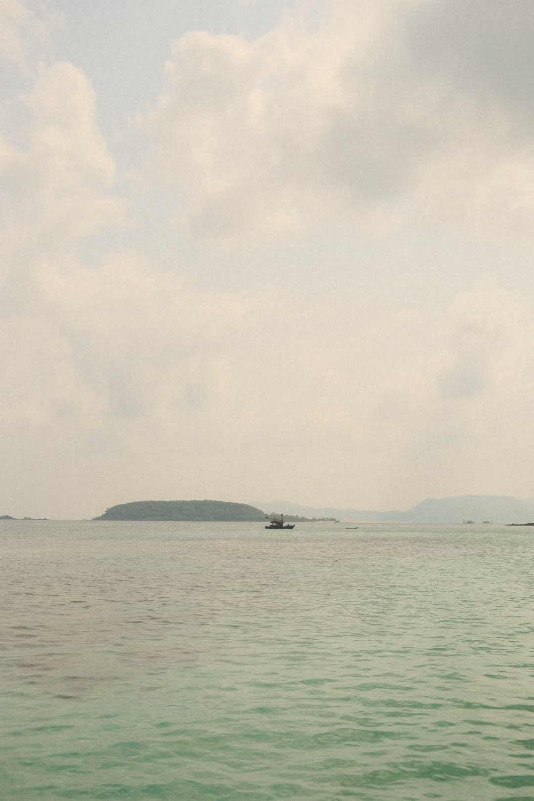 A low, flat green island on the horizon with a small boat, calm green-grey sea and hazy sky.