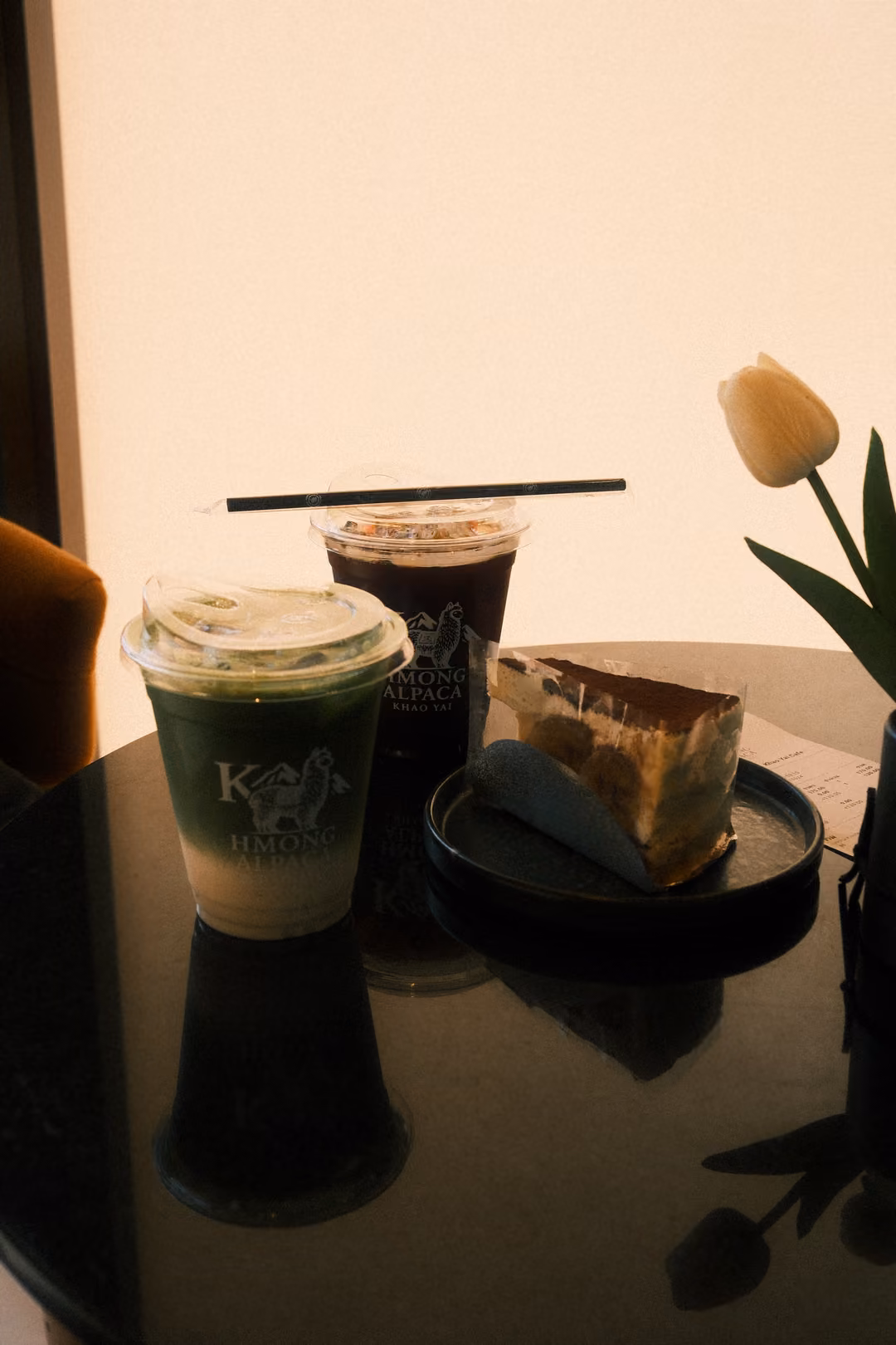 The same table with the green drink, iced coffee, cake and tulip, all reflected in the dark surface.