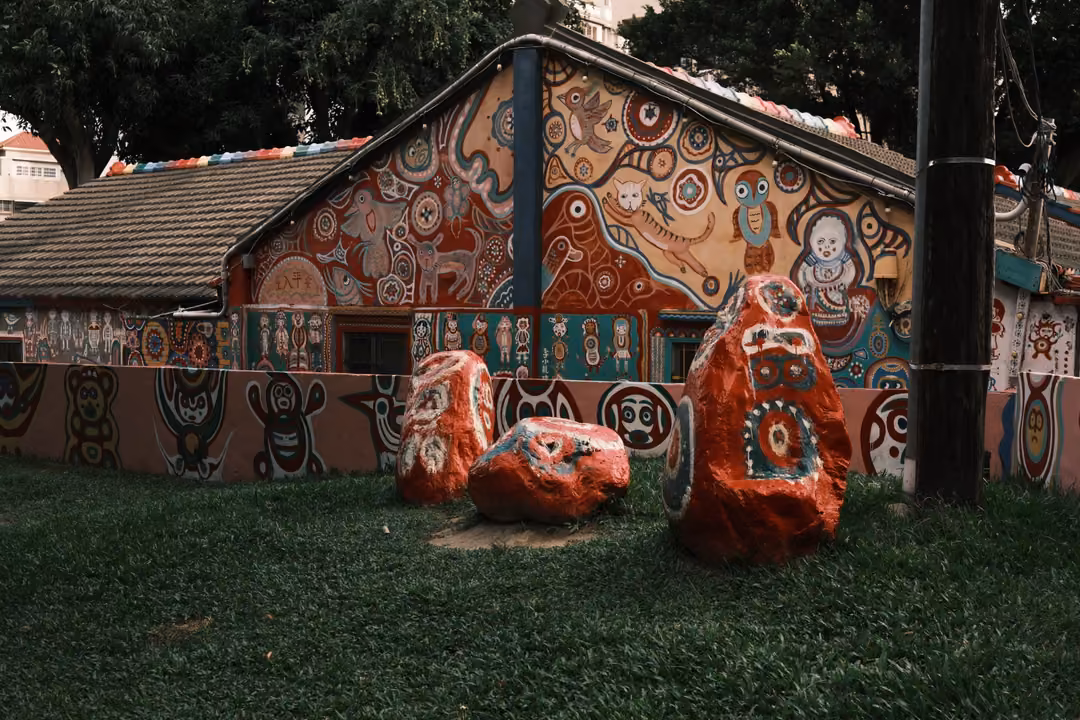 Painted house gable covered in colourful figures, red and orange painted boulders on a lawn in front