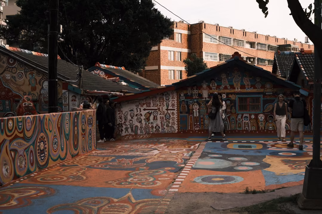Painted courtyard and low houses covered in colourful figures, visitors walking, apartment blocks behind