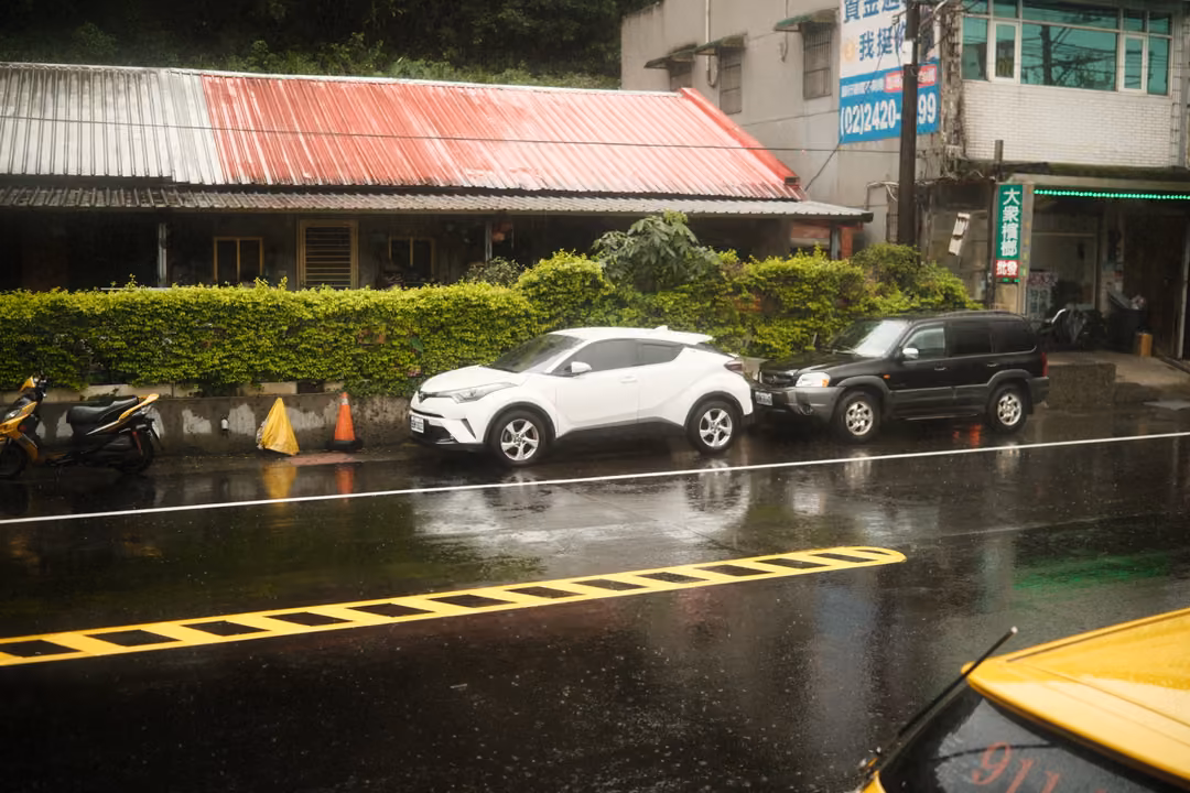Same rainy roadside with parked white and black cars and a rust-red roof, framed slightly differently