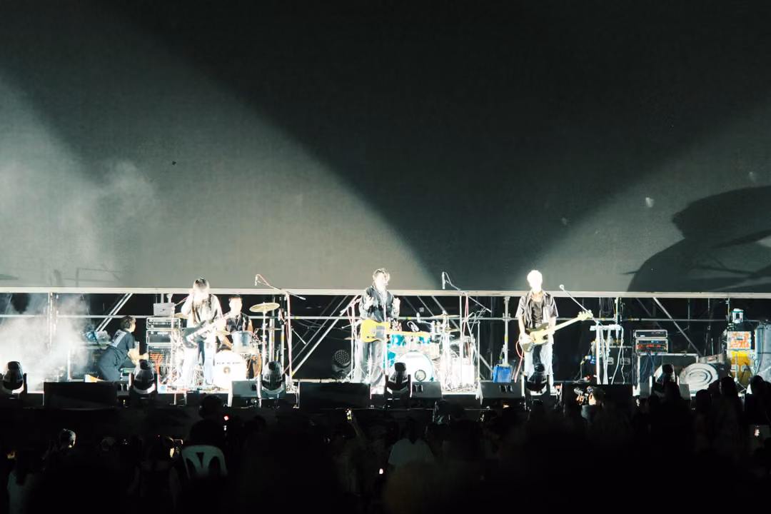 Band on a wide stage under pale searchlight beams and white haze, silhouetted crowd and chairs in front