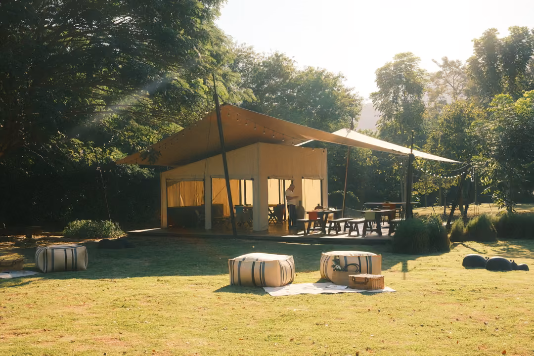 Canvas-roofed pavilion with sunbeams falling across a lawn of striped poufs, trees and hills behind