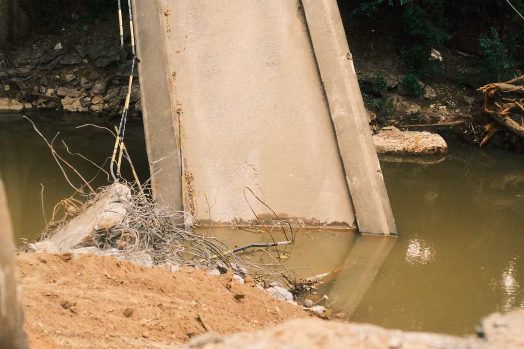 A collapsed concrete bridge slab sloping down into murky brown river water, dirt and debris on the bank.