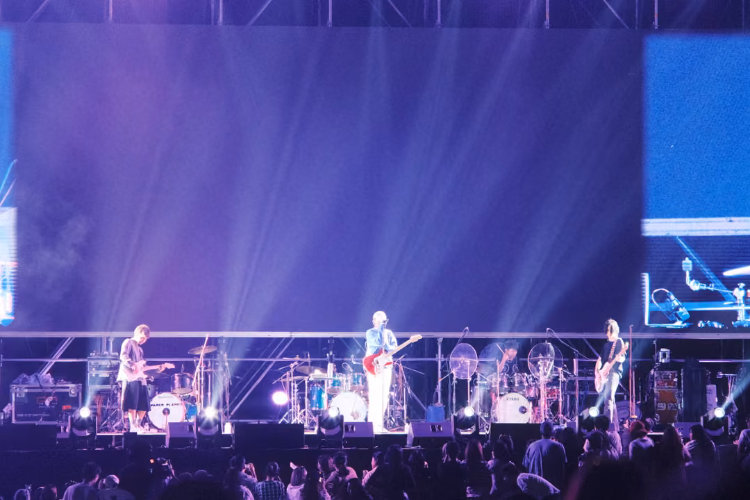 Band performing on a purple stage beneath an empty violet screen, beams of white light rising through haze.