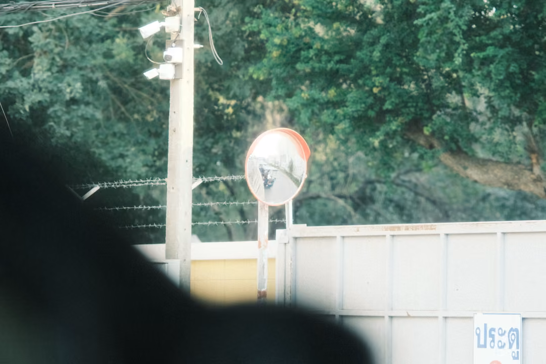 A convex traffic mirror on a post beside a white wall and gate, with security cameras above and a dark blur in front.