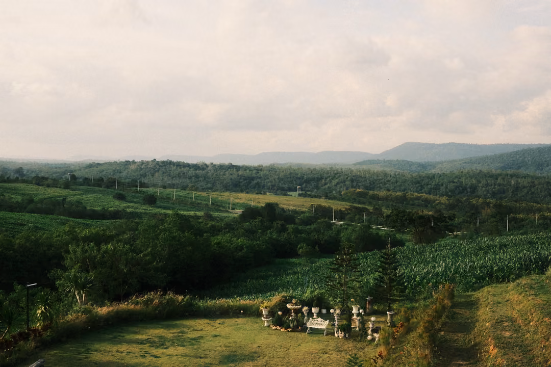 A green valley of fields and forest below a cloudy sky, a lawn with white garden statues in the foreground.