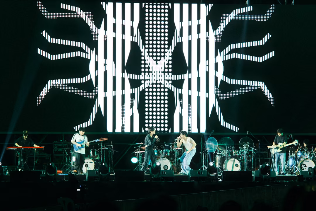 Screen with dense white vertical stripes over a branching dotted pattern, band performing on stage below.