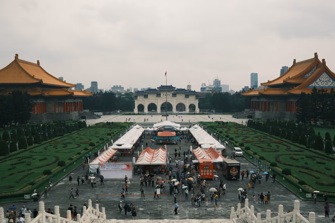 Brighter view of the plaza with white festival tents, crowds and umbrellas between two orange-roofed halls