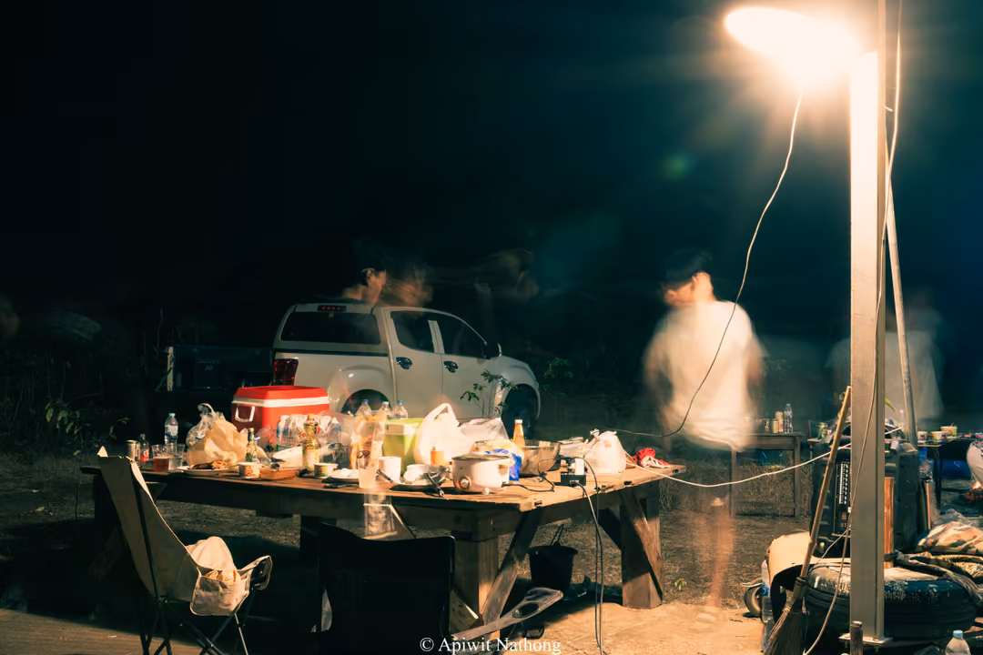 Camp table loaded with food, bottles and a cooler under a bright lamp, a pickup truck and a blurred figure behind