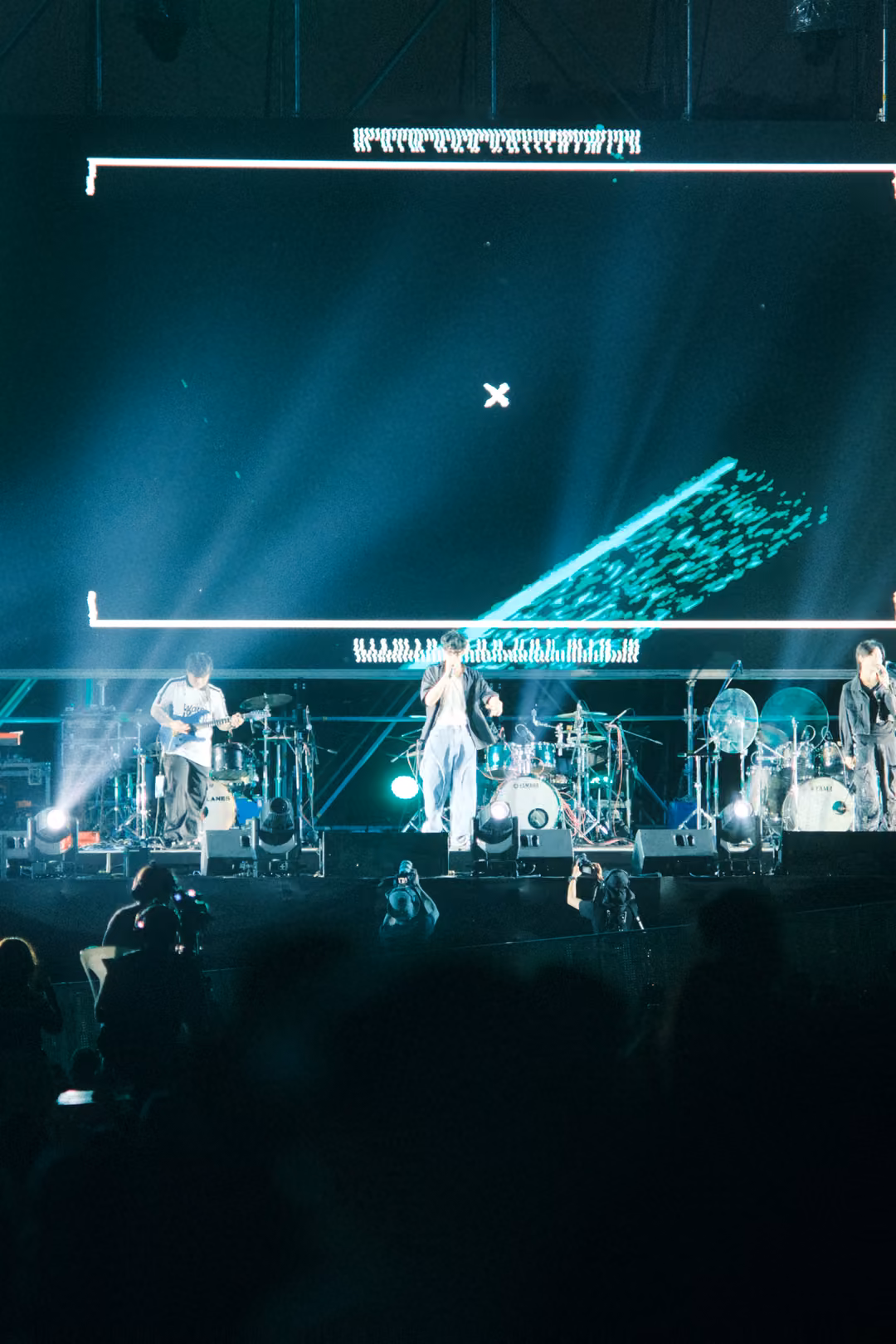 Vertical view of a stage with white light beams cutting through haze toward the crowd, a turquoise slab on screen.
