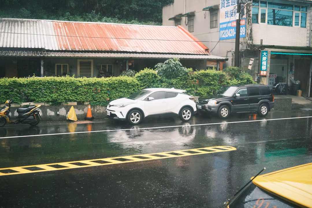 Rain-soaked street with parked cars beside a hedge and rusted roof, a yellow bonnet and wiper in the corner