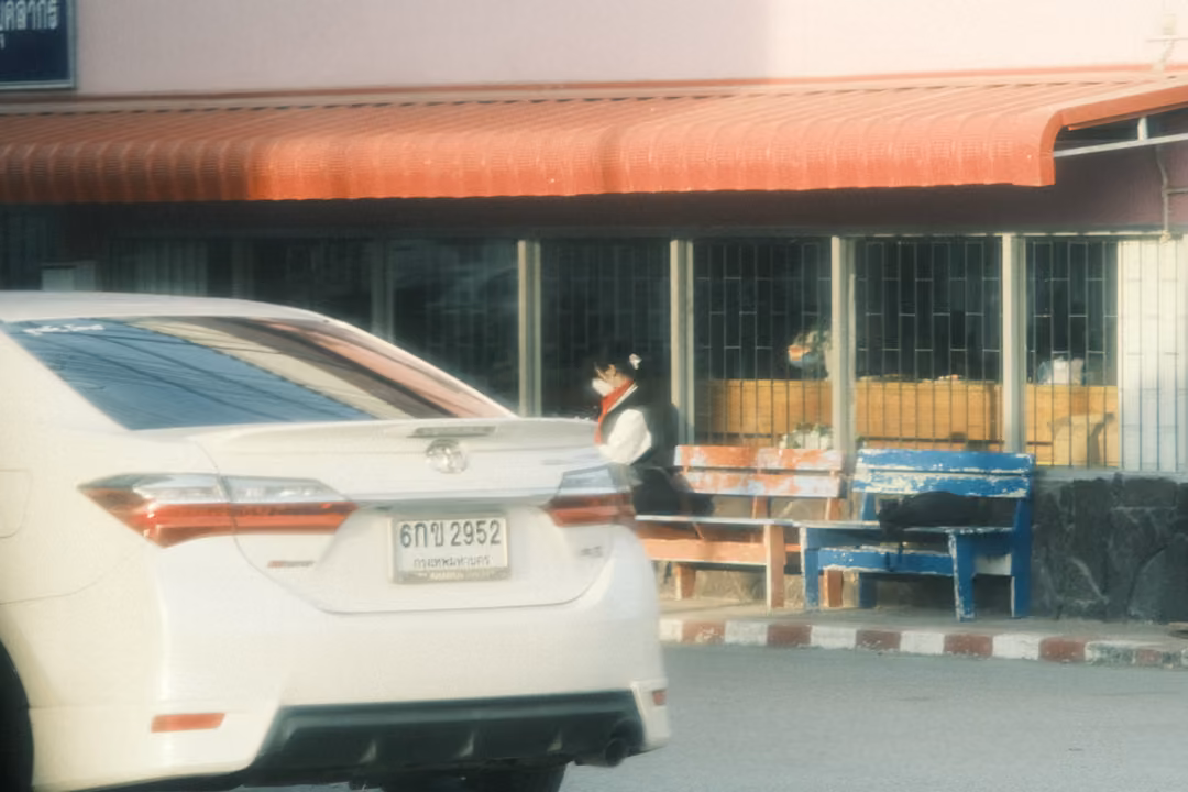 The rear of a white sedan parked before a building with a red awning, a masked student seated on a blue bench.